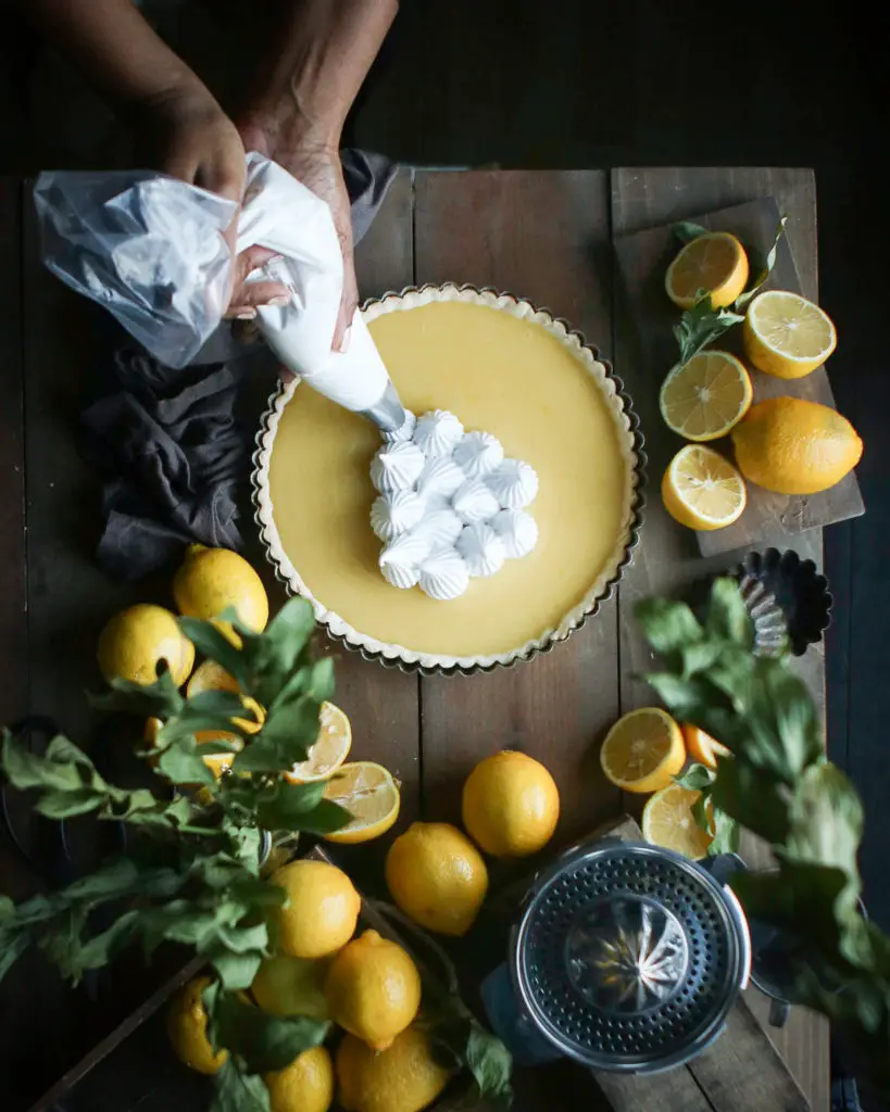 Large lemon tart on a brown wooden table. a Black women is piping dollops of white egg white based meringue dolloped on top. Lemons, a citrus juicer and green leaves are used as decorations around the tart.