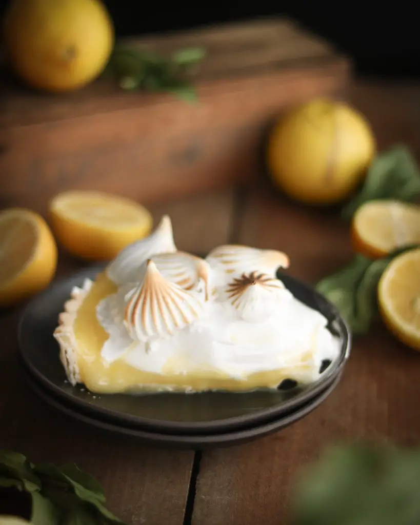 A piece of lemon tart with white egg white based meringue dolloped on top. The meringue is slightly browned from a torch. Lemons and green leaves are used as decorations around the tart.