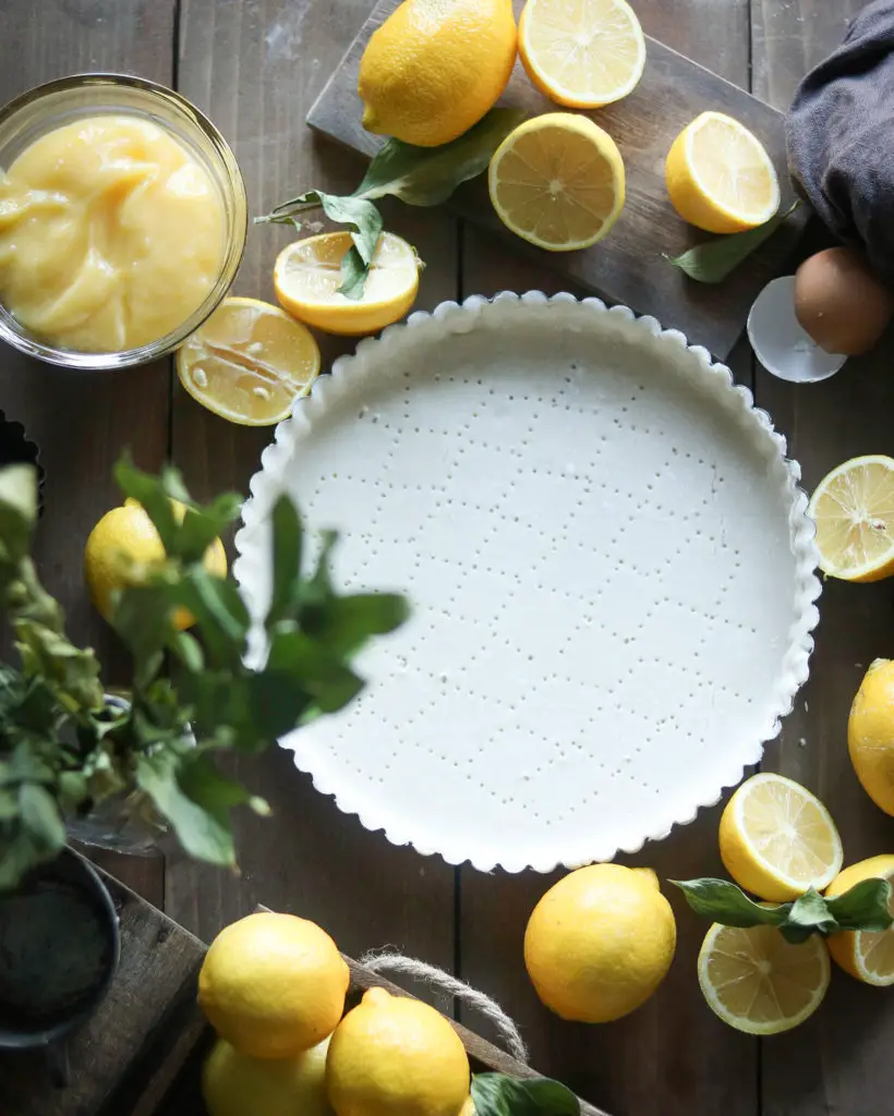 Large pastry tart shell is sitting on a brown wooden table. Lemons, lemon pie filling in a clear bowl, and green leaves are used as decorations around the tart.