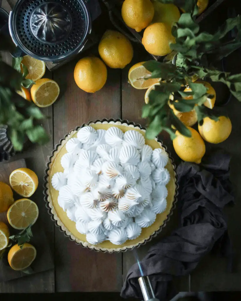 Large lemon tart with white egg white based meringue dolloped on top. The meringue is being slightly browned from a torch. Lemons and green leaves are used as decorations around the tart.