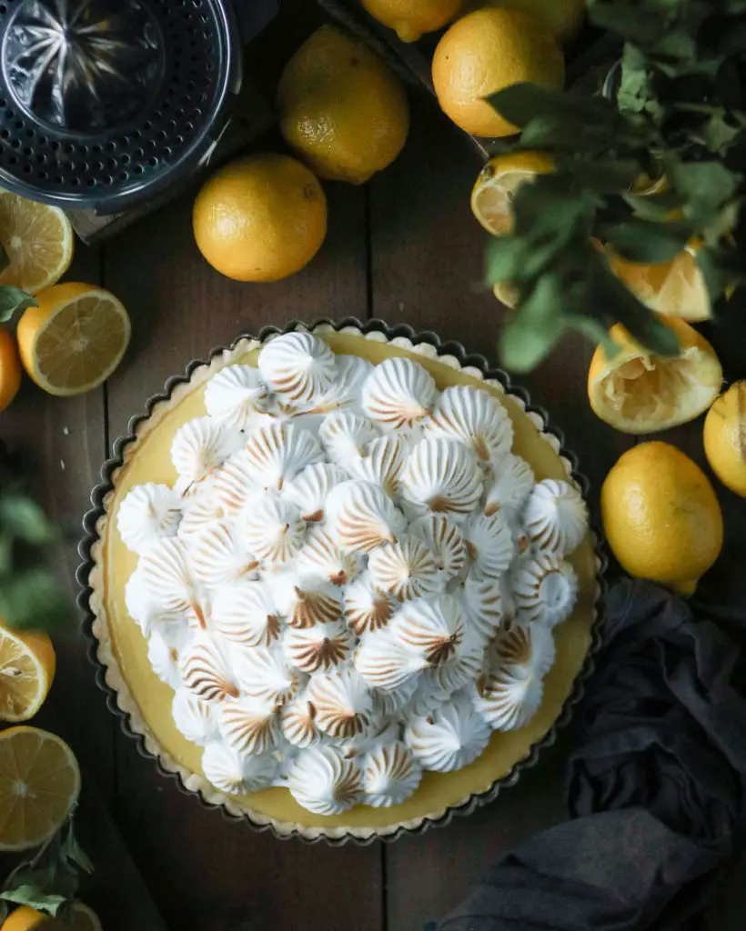 Large lemon tart with white egg white based meringue dolloped on top. The meringue is slightly browned from a torch. Lemons and green leaves are used as decorations around the tart.