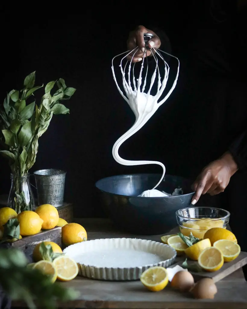 Large pastry tart shell is on a wooden table. A Black women is holding a whisk attachment from a stand mixer with meringue coming down from it in a wave movement. Lemons, egg shells, a metal cup and green leaves are used as decorations around the tart.