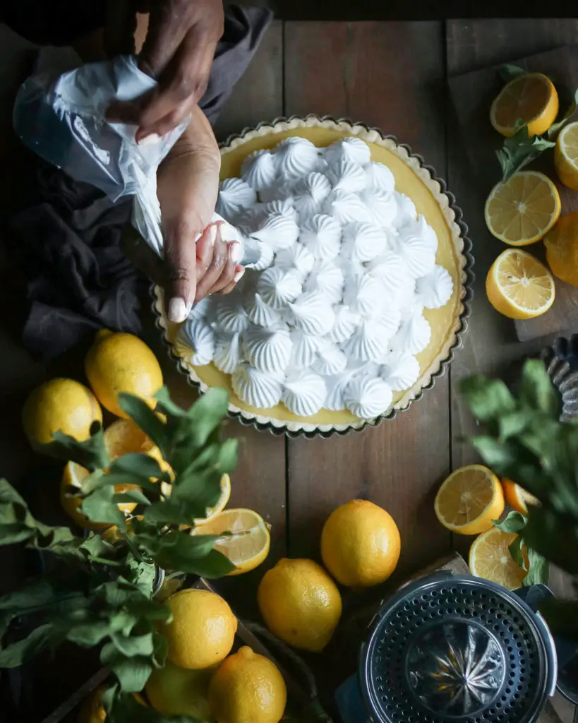 Large lemon tart on a brown wooden table. A Black women is piping dollops of white egg white based meringue dolloped on top. Lemons, a citrus juicer and green leaves are used as decorations around the tart.