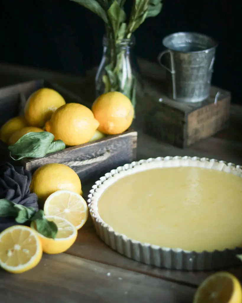 Large plain lemon tart is sitting on a wooden table. Lemons, a metal cup and green leaves are used as decorations around the tart.