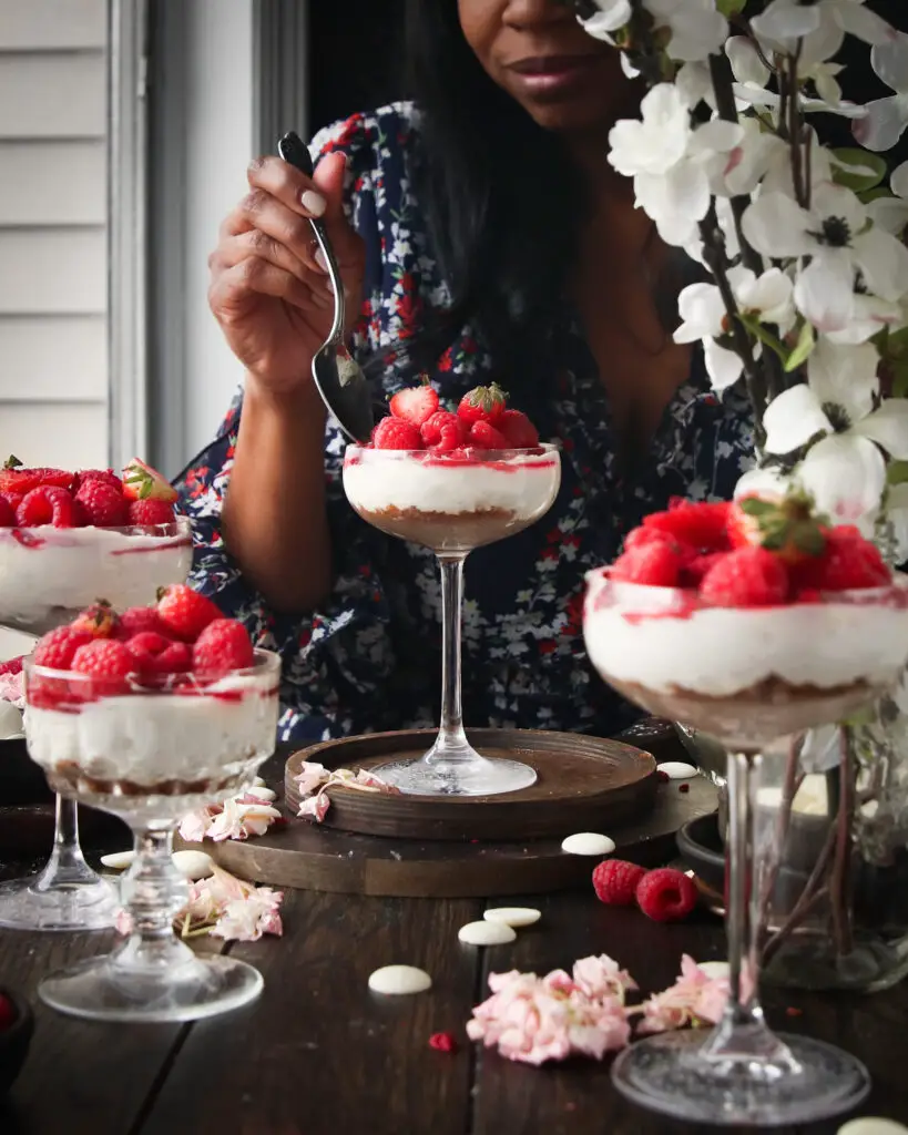 A Black women eating a coupe glass filled with cheesecake filling and berries. Strawberries, white chocolate pieces, and pink and white flowers are in the background.