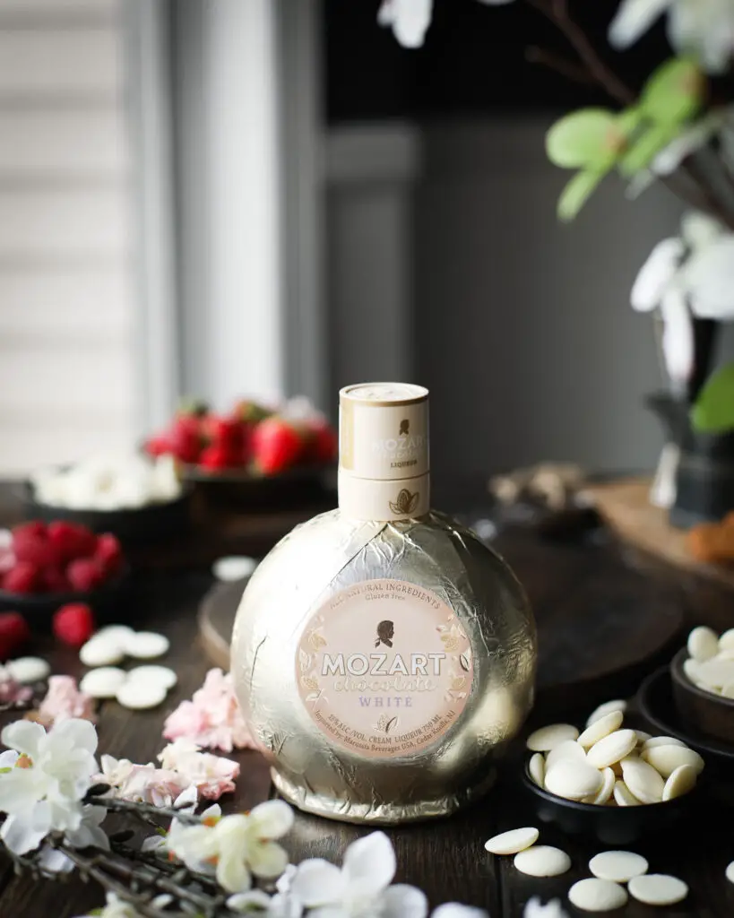 Bottle of Mozart White Chocolate Liqueur. Berries, white chocolate pieces and flowers are in the background.