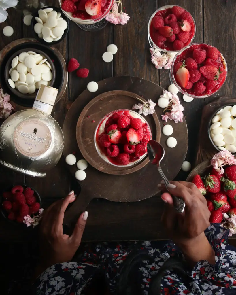 An overhead picture of a black women sitting at a wooden table eating coupe glasses filled with cheesecake filling topped with berries and crushed freeze dried berries. Pink and white flowers, berries, white chocolate pieces and a Mozart white chocolate liqueur bottle are in the background.