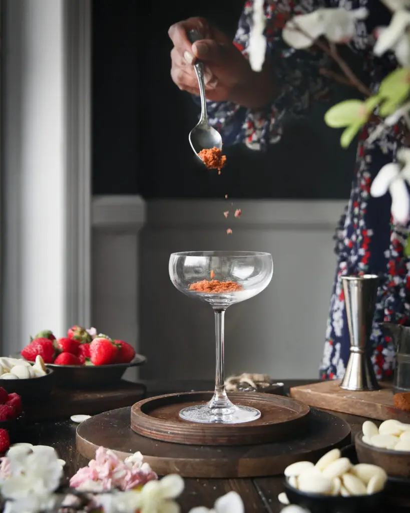 A Black women sprinkling crushed cookies into a coupe glass. Strawberries, white chocolate pieces and pink and whites flowers are in the background.