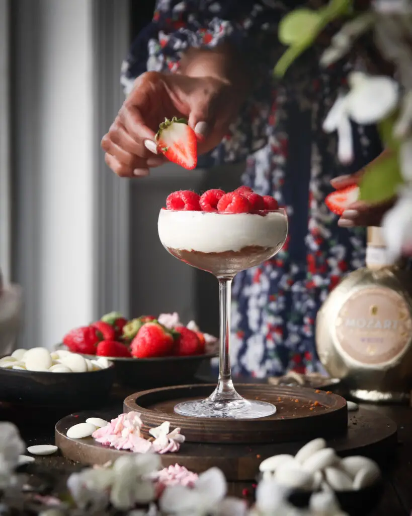 A Black women adding berries to a coupe glass filled with cheesecake filling. Strawberries, white chocolate pieces, a Mozart liqueur bottle and pink and white flowers are in the background.