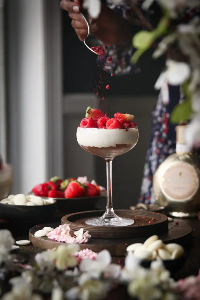 A Black women adding freeze dried berries to a coupe glass filled with cheesecake filling and berries. Strawberries, white chocolate pieces, a Mozart liqueur bottle and pink and white flowers are in the background.