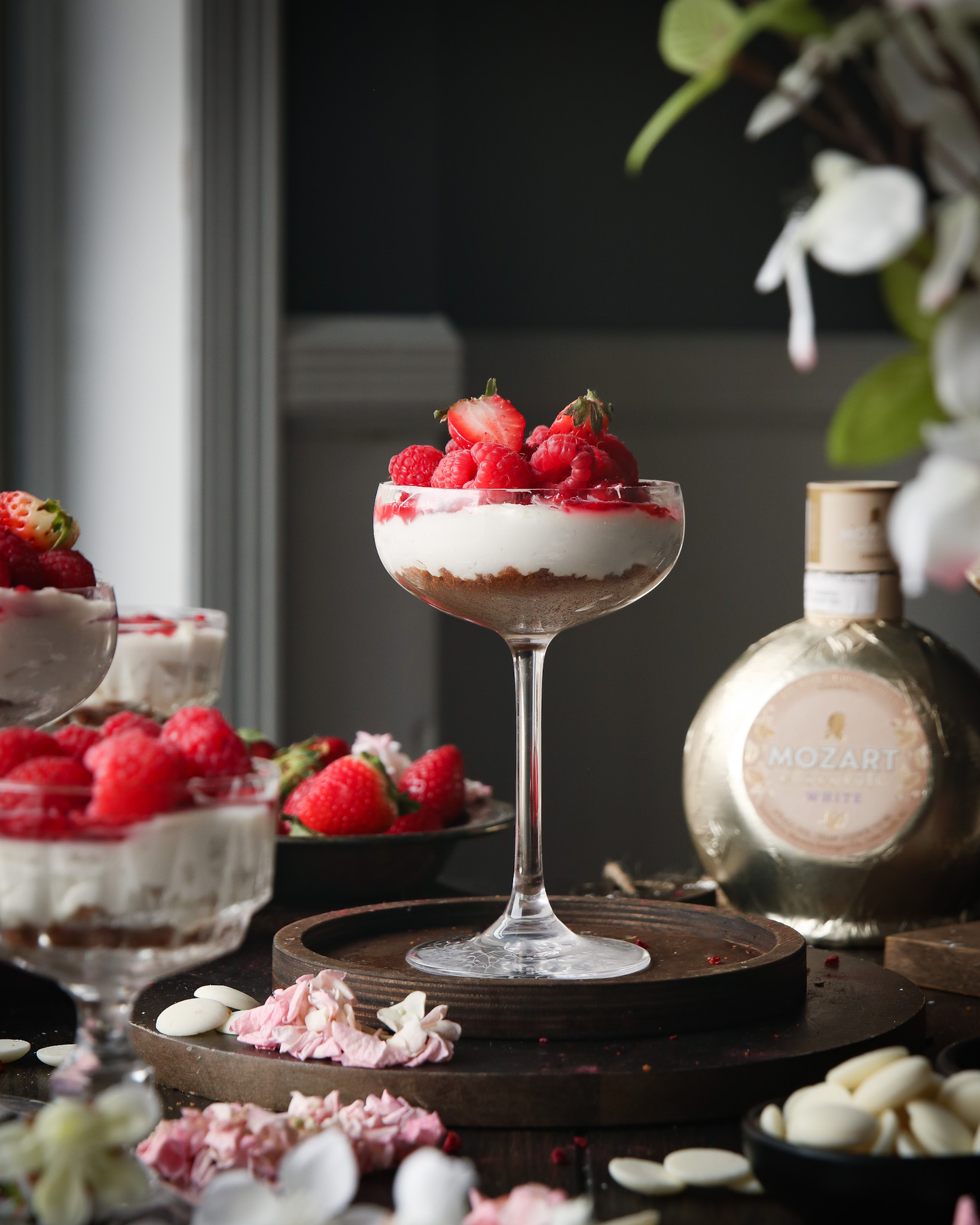 A coupe glass filled with cheesecake filling and berries. Strawberries, white chocolate pieces, a Mozart liqueur bottle, and pink and white flowers are in the background.