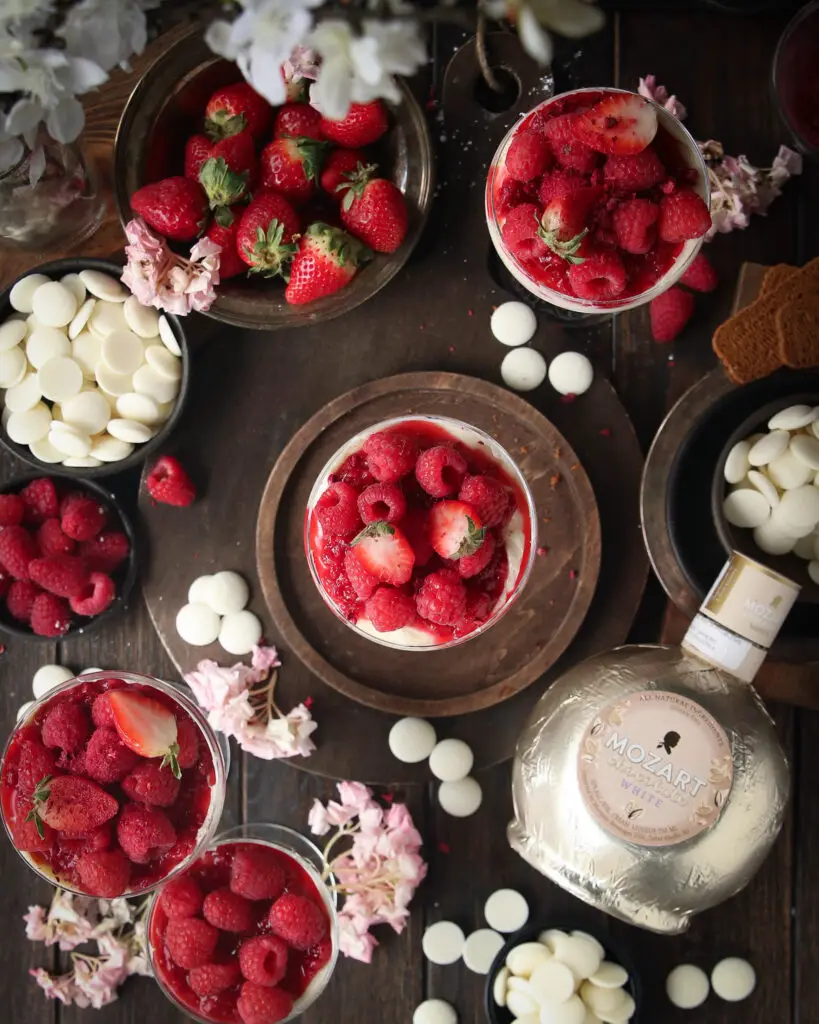 An overhead picture of coupe glasses filled with cheesecake filling topped with berries and crushed freeze dried berries. Pink and white flowers, berries, white chocolate pieces and a Mozart white chocolate liqueur bottle are in the background.