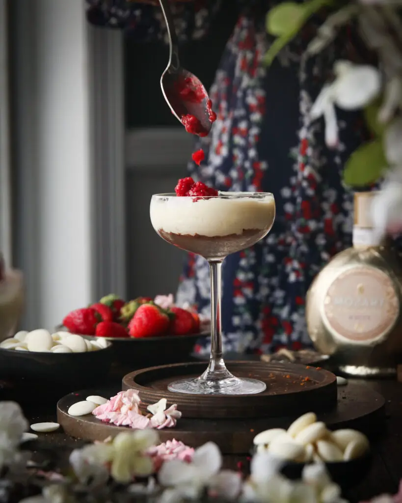 A Black women adding berry filling to a coupe glass filled with cheesecake filling. Strawberries, white chocolate pieces, a Mozart liqueur bottle and pink and white flowers are in the background.