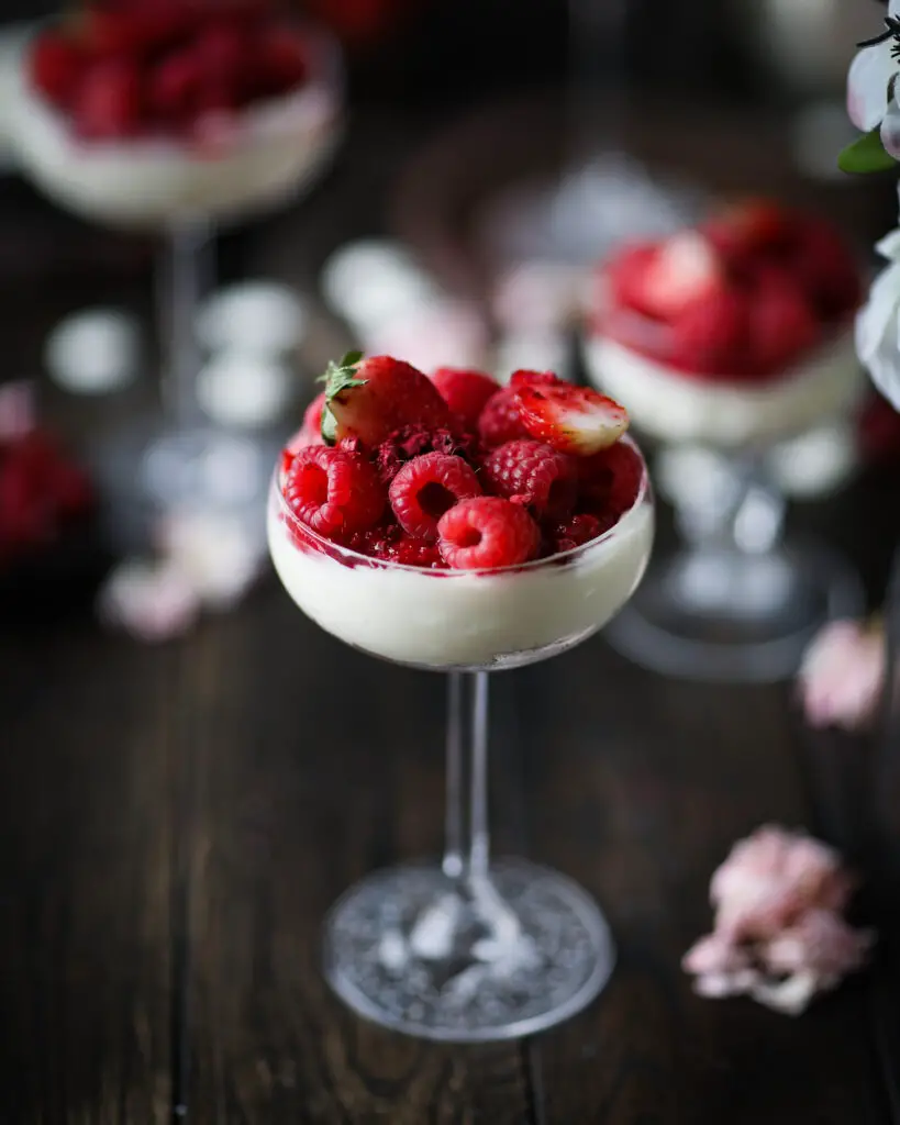 Coupe glass filled with cheesecake filling topped with berries and crushed freeze dried berries. Additional glasses and pink flowers are in the background.