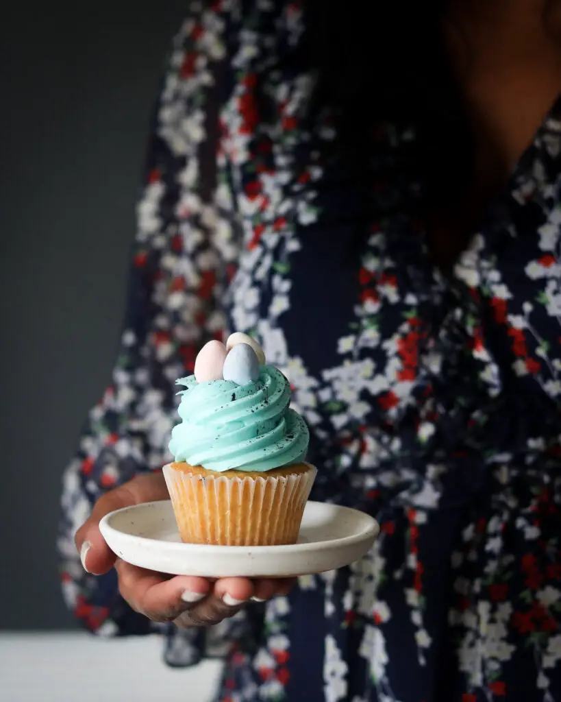 A Black woman holding a vanilla cupcake topped with a swirl of blue buttercream, splattered with black food coloring and 3 candy easter eggs on top. The cupcake it sitting on a small beige plate.