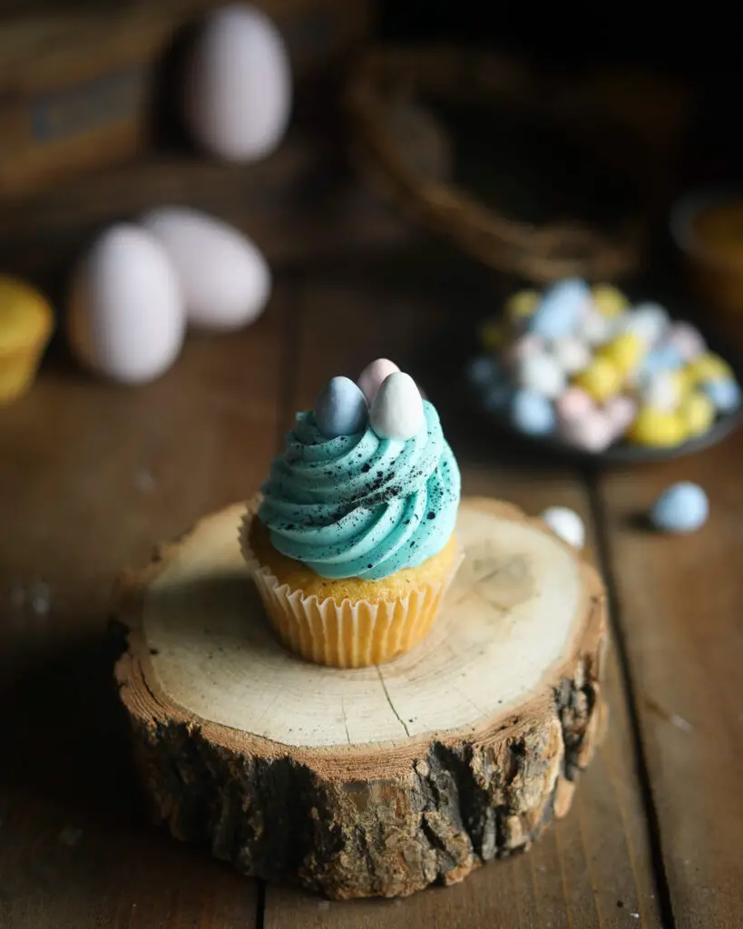 A vanilla cupcake topped with a swirl of blue buttercream, splattered with black food coloring and 3 candy easter eggs on top. The cupcake it sitting on a wooden trunk piece. easter eggs in the background. 