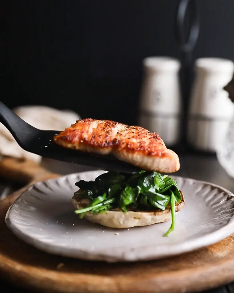 English muffin topped with spinach. A piece of salmon is being placed on top with a spatula.  It is on a tan plate on a round wooden board. 