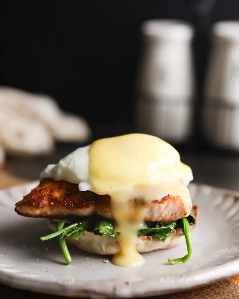 English muffin topped with spinach, a piece of salmon, a poached egg, and a drizzle of yellow hollandaise sauce. It is on a tan plate on a round wooden board. 