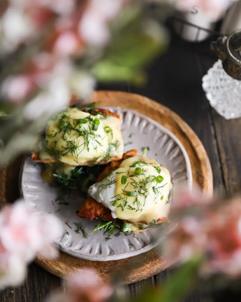 An overhead look at an English muffin topped with spinach, a piece of salmon, a poached egg, a drizzle of yellow hollandaise sauce and a sprinkle or chopped dill and green onions. It is on a tan plate on a round wooden board. The plate is on a wooden table. Pink flowers out of focus at the forefront of the photo.