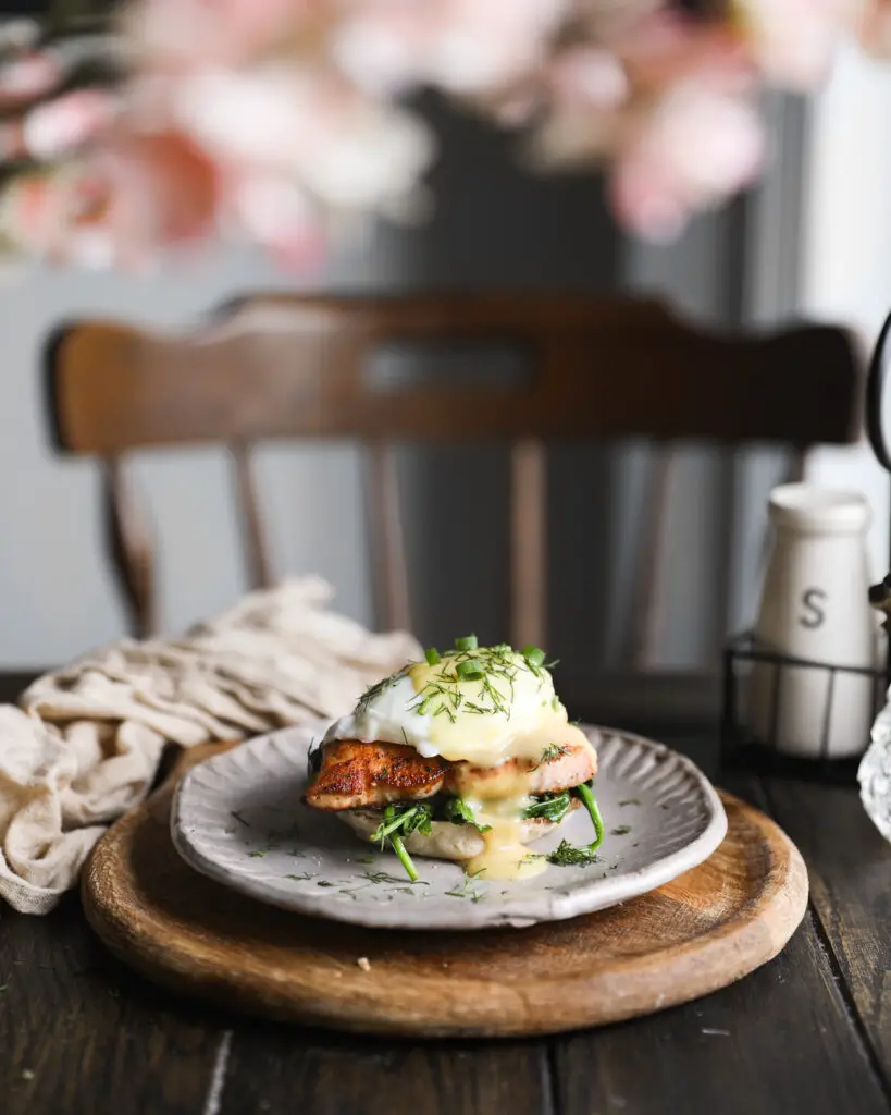 English muffin topped with spinach, a piece of salmon, a poached egg, a drizzle of yellow hollandaise sauce and a sprinkle or chopped dill and green onions. It is on a tan plate on a round wooden board. The plate is on a wooden table with a chair in the background, and pink flowers out of focus at the top of the photo.