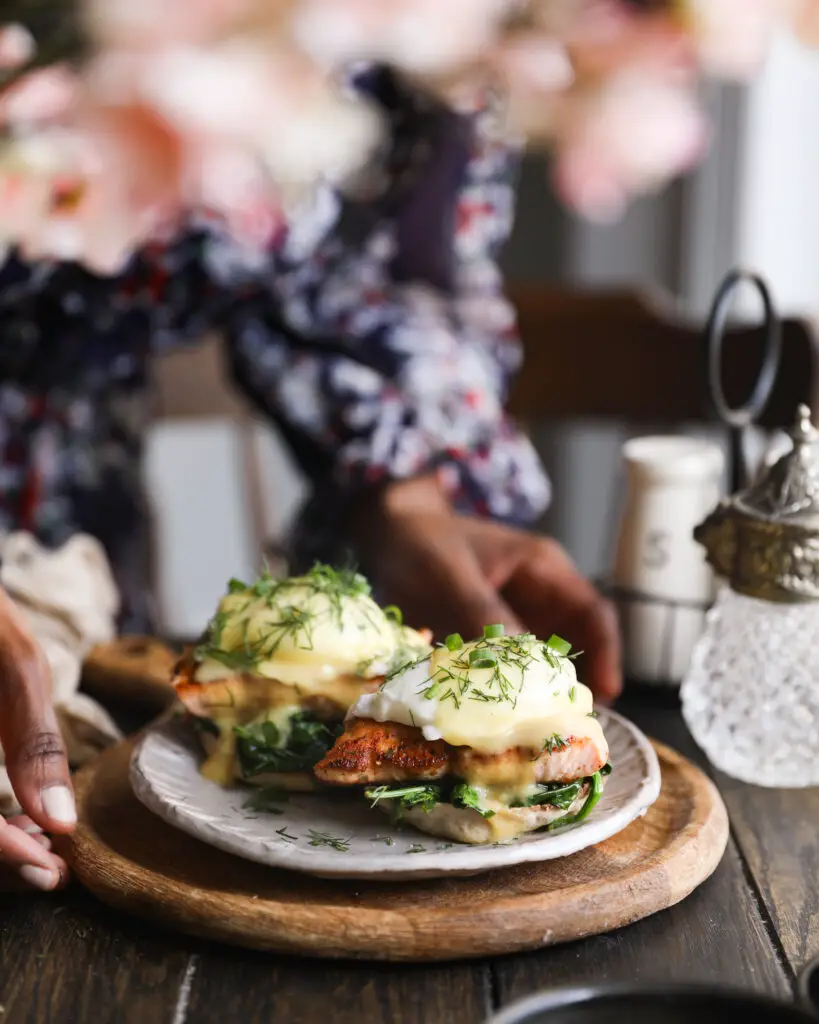 English muffin topped with spinach, a piece of salmon, a poached egg, a drizzle of yellow hollandaise sauce and a sprinkle or chopped dill and green onions. It is on a tan plate on a round wooden board. A Black woman is holding the plate. The plate is on a wooden table with a chair in the background, and pink flowers out of focus at the top of the photo.