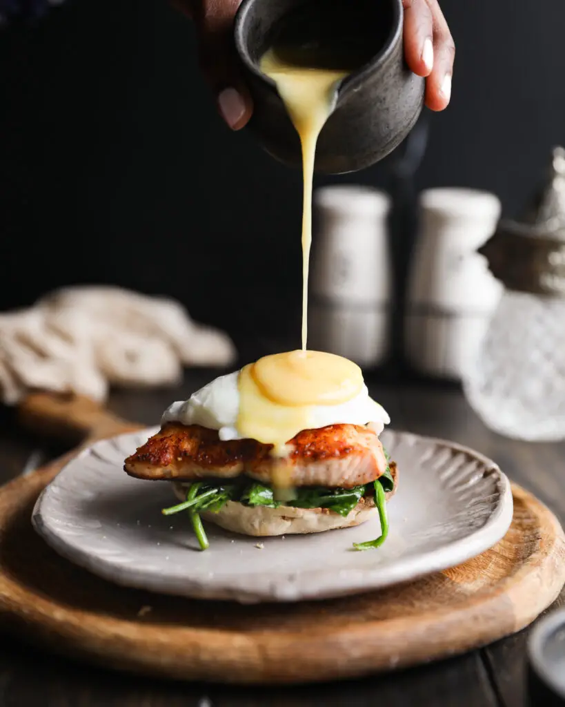 English muffin topped with spinach, a piece of salmon, and a poached egg. A hand is pouring on yellow hollandaise sauce from a cup. It is on a tan plate on a round wooden board. 