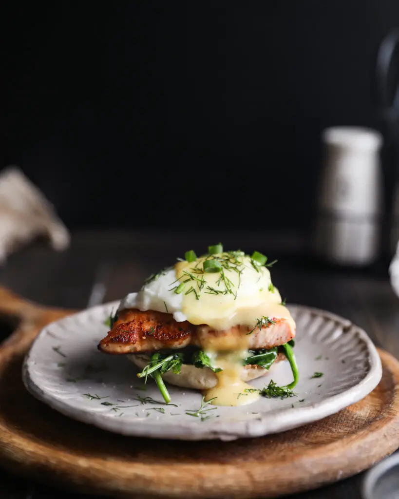 English muffin topped with spinach, a piece of salmon, a poached egg, a drizzle of yellow hollandaise sauce and a sprinkle or chopped dill and green onions. It is on a tan plate on a round wooden board. 