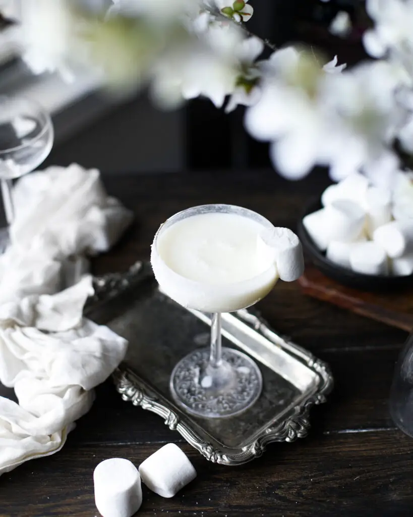 White creamy cocktail on a silver tray with a marshmallow garnish. White flowers are highlighted out of focus at the top of the picture.