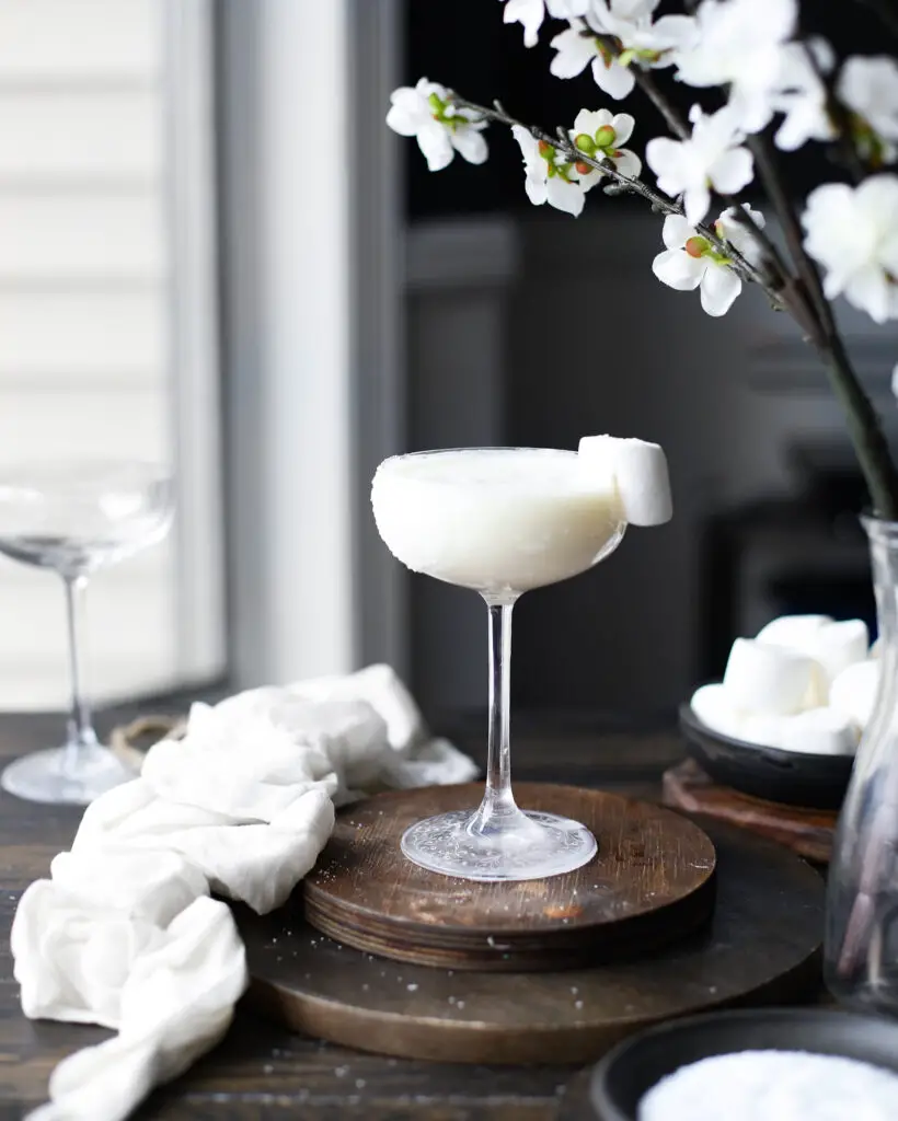 White creamy cocktail on a round wooden coaster with a marshmallow garnish. White flowers are highlighted out of focus at the top of the picture.