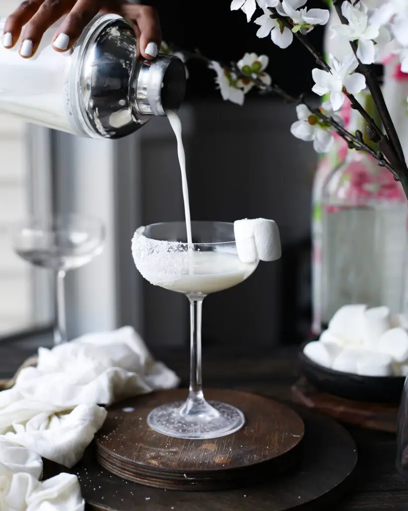 Black woman pouring a white creamy cocktail into a coupe glass on a round wooden coaster with a marshmallow garnish. White flowers are highlighted out of focus at the top of the picture. 