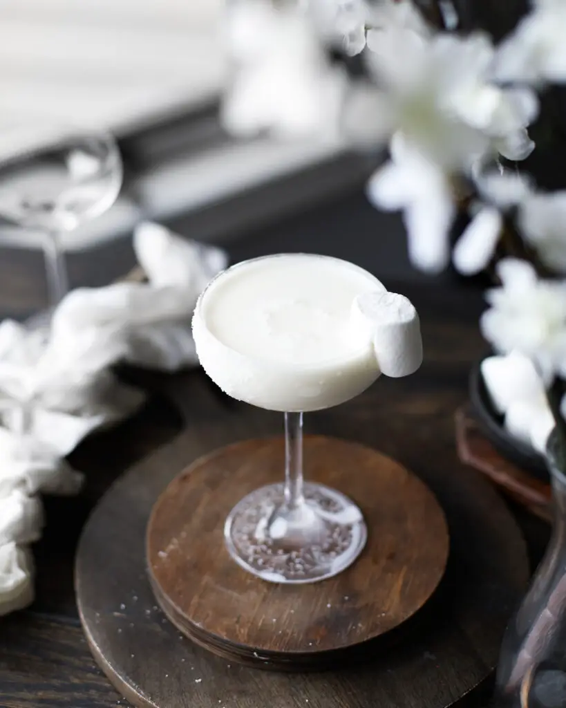 White creamy cocktail on a round wooden coaster with a marshmallow garnish. White flowers are highlighted out of focus at the top of the picture.